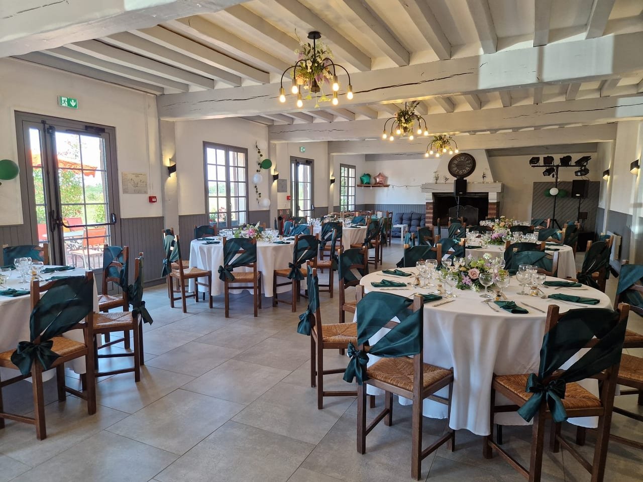 La salle en enfilade, tables rondes dressées, cheminée au fond et portes-fenêtres ouvertes sur la terrasse