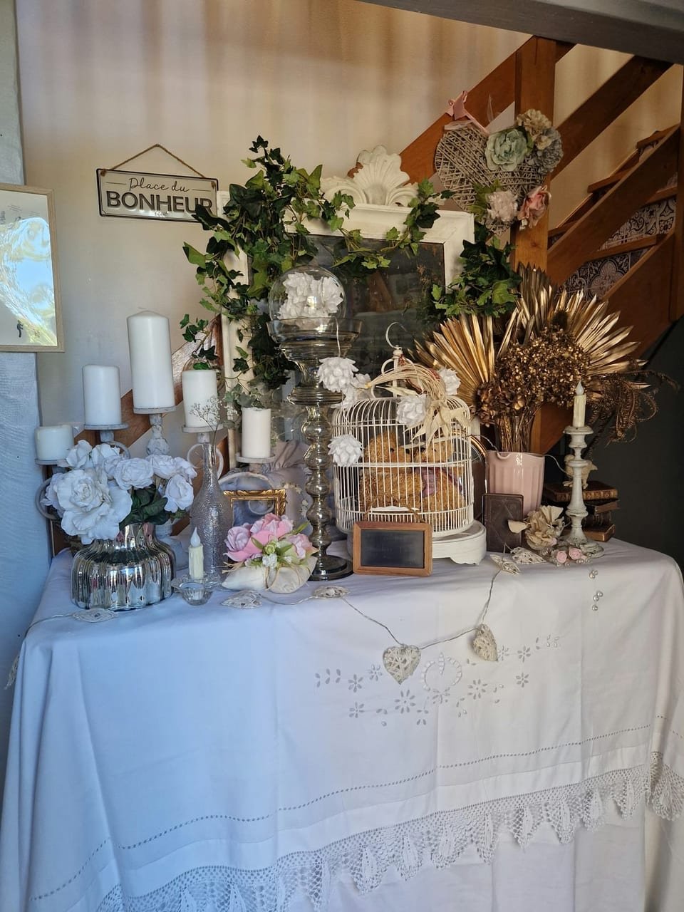 Table de décoration sous l’escalier : bougies, cage blanche, fleurs séchées et miroir ancien